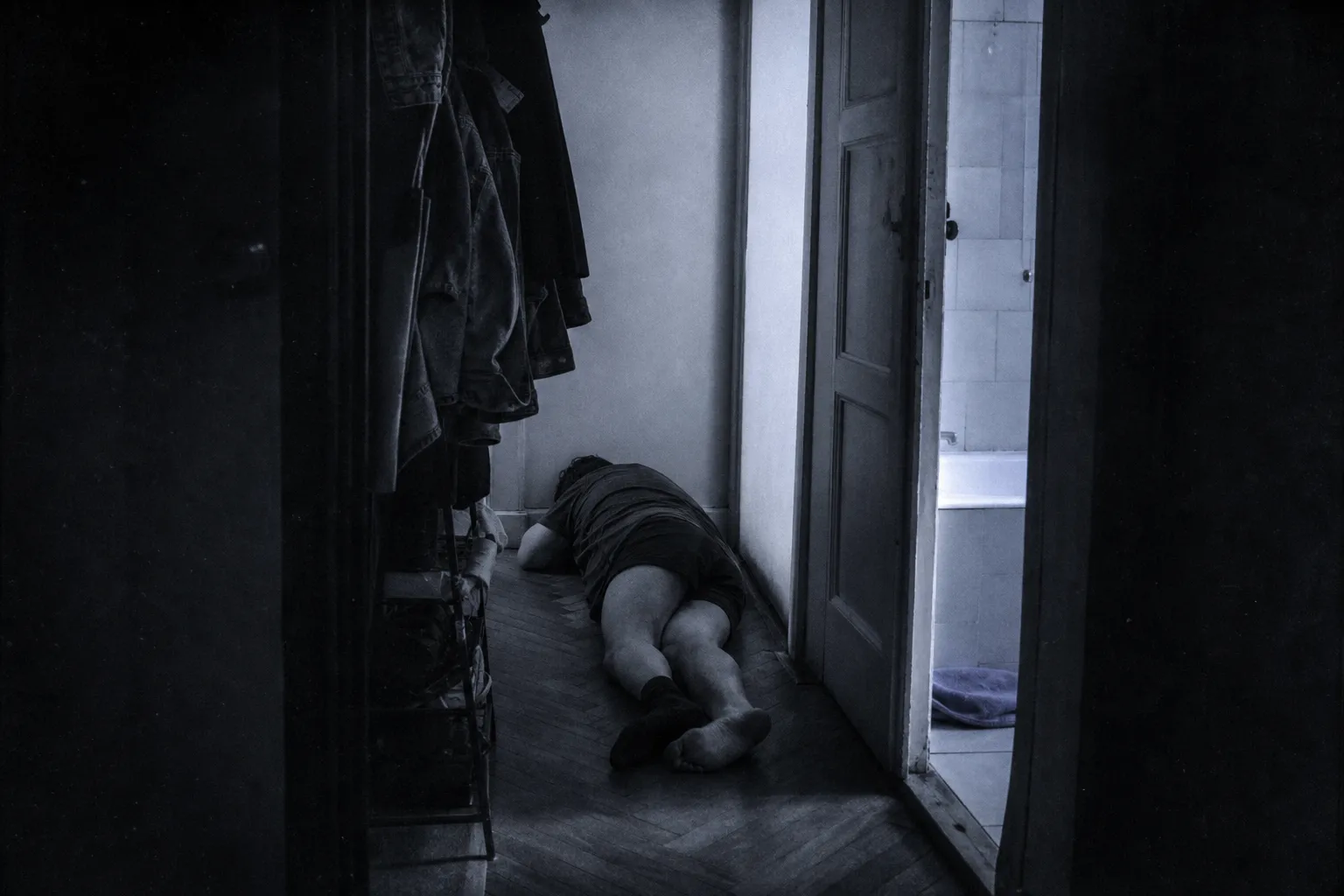 The narrow hallway of a small flat in grey morning light: a man asleep on the floor between the coat rack and the bathroom door, in shorts and one sock, face turned away.