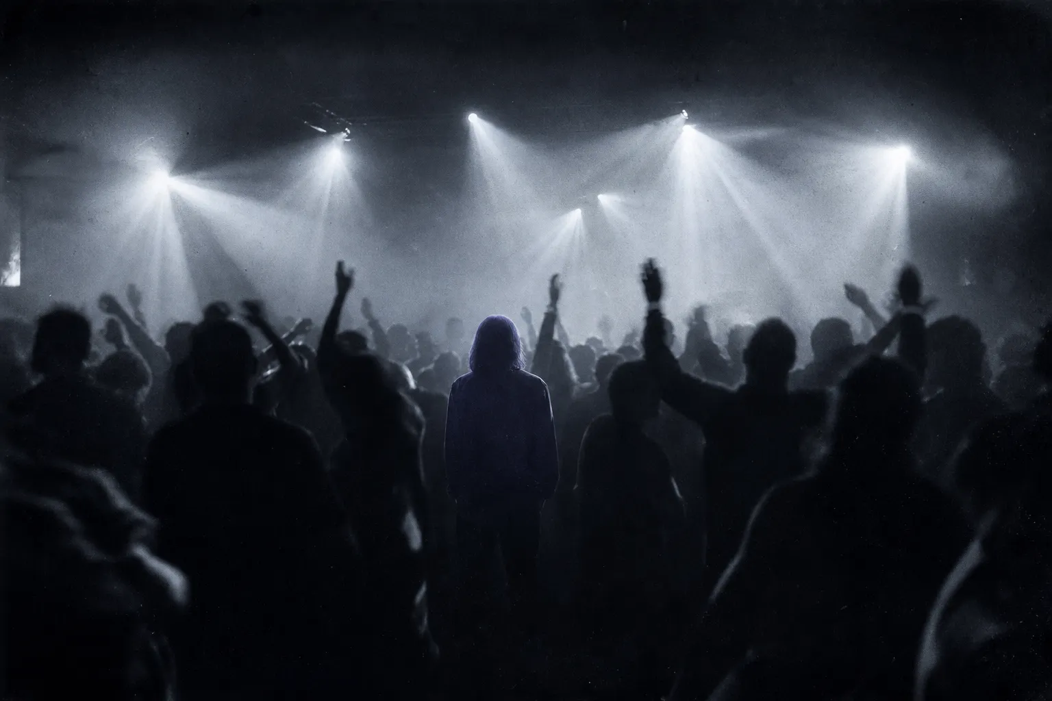 A late-1990s trance party in a warehouse: a crowd of dancing silhouettes, arms raised, light beams in smoke, one figure standing still in the middle.