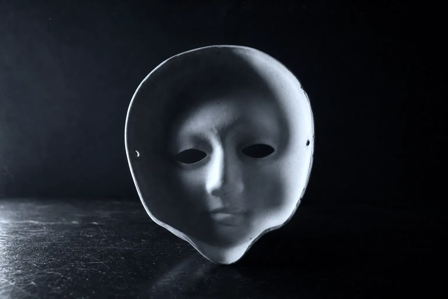 A white carnival mask turned inside-out toward us, yet it reads as a convex face emerging from shadow.