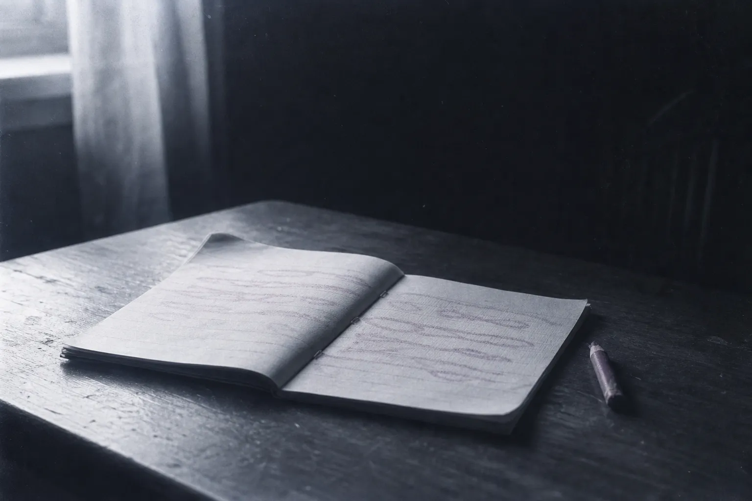 A child's notebook open on a wooden table, a few lines written large in red pencil, the worn-down red pencil beside it; spring light from a window.